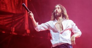 A man with long hair and a beard, wearing an open white shirt and black pants, holds a microphone extended in one hand on a red-lit stage.
