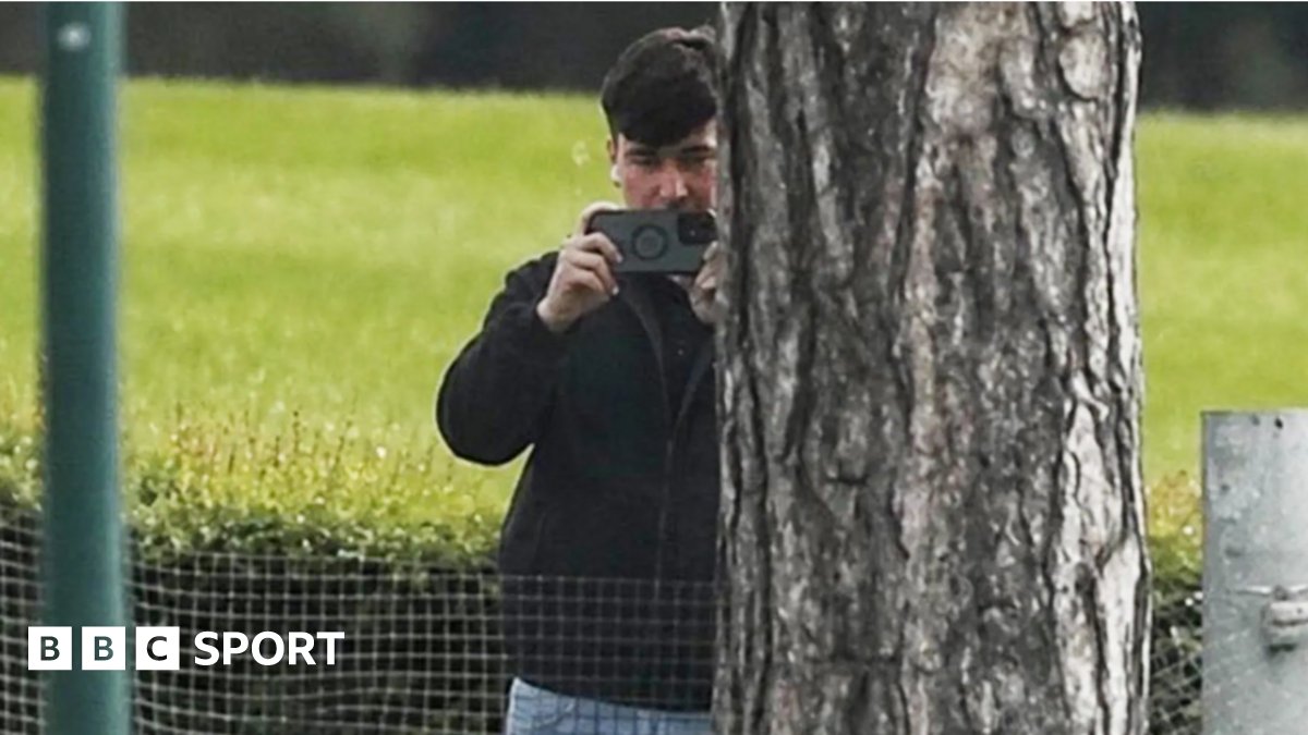 A man in a dark jacket and blue jeans stands partially concealed by a tree trunk and holds a phone up in two hands as if filming and has a white earphone in his right ear